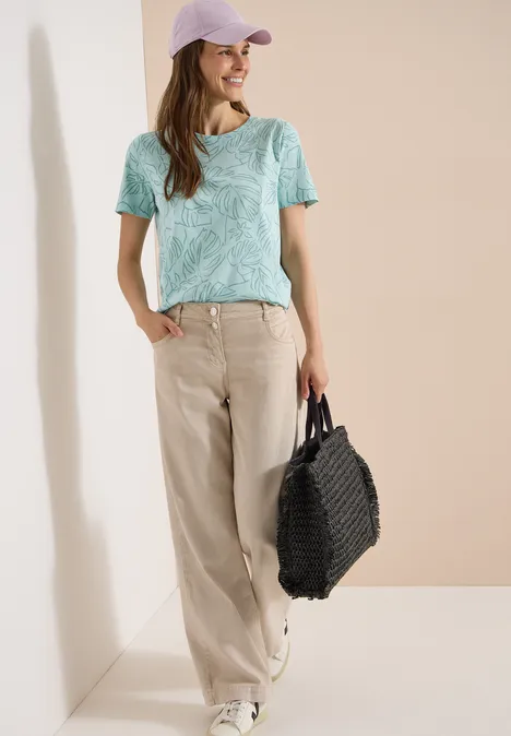 T-Shirt mit Rundhals und Blätterprint crystal mint Frau lehnt an weißer Wand in mintgrünem Shirt, beigen weiten Hosen, weißen Sneakern, schwarzen Tasche und rosa Cap.