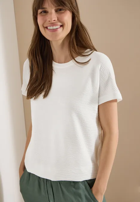 Frau trägt weißes Kurzarmshirt und grüne Hose, Hände in den Taschen vor neutralem Hintergrund.