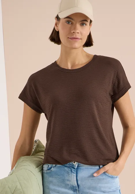 Vrouw in bruin T-shirt en blauwe jeans leunt ontspannen tegen een groen kussen voor een beige achtergrond.