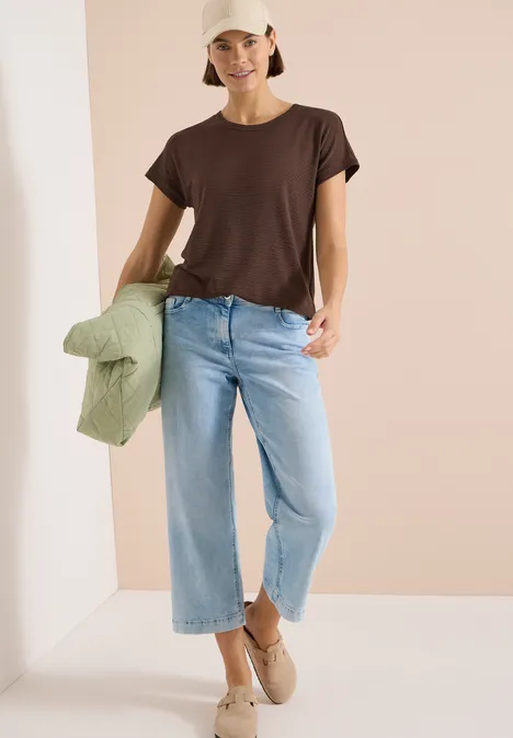 Person draagt bruin T-shirt, lichte jeans en beige schoenen, houdt een groen jack in de hand.