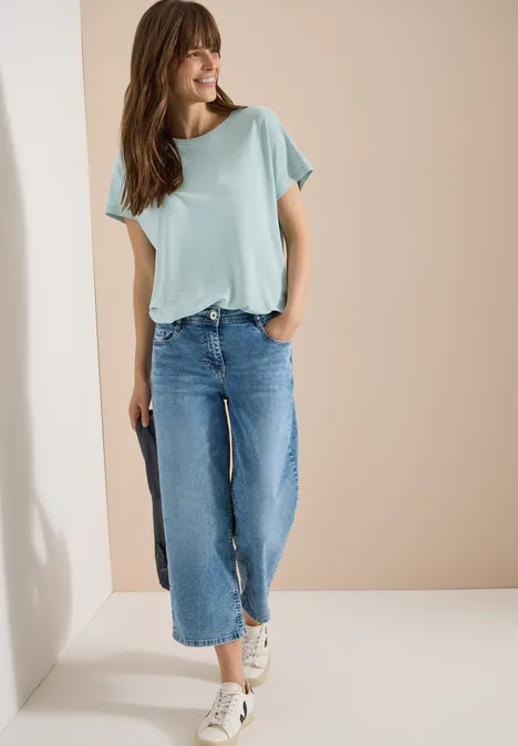 T-Shirt mit Rundhals und Struktur crystal mint Frau in mintgrüner Bluse und weit geschnittener, kurzer Jeanshose lächelnd vor beigem Hintergrund.