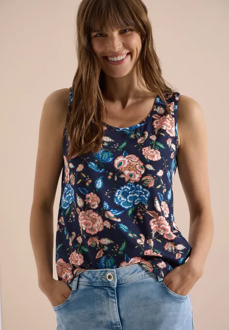 Frau trägt ärmelloses blaues Oberteil mit Blumenmuster und Jeans, Hände in den Taschen.