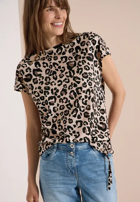 Frau in Leopardenmuster-T-Shirt mit seitlichen Bändern kombiniert mit blauer Jeans vor neutralem Hintergrund.