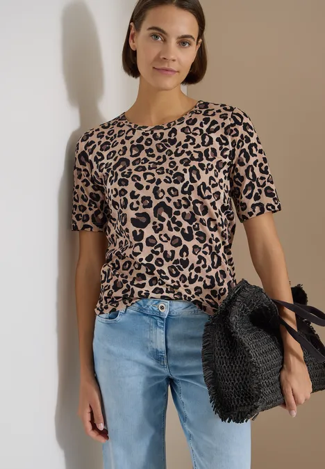 Person in beige-schwarzem Leoparden-Shirt, blauen Jeans und schwarz geflochtener Tasche vor neutralem Hintergrund.