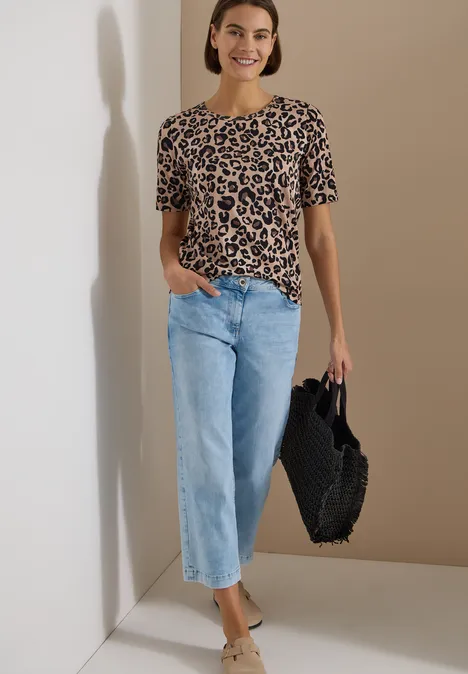 Frau in Leoparden-Shirt, hellblauen Jeans und schwarzer Flecht-Handtasche vor neutralem Hintergrund.