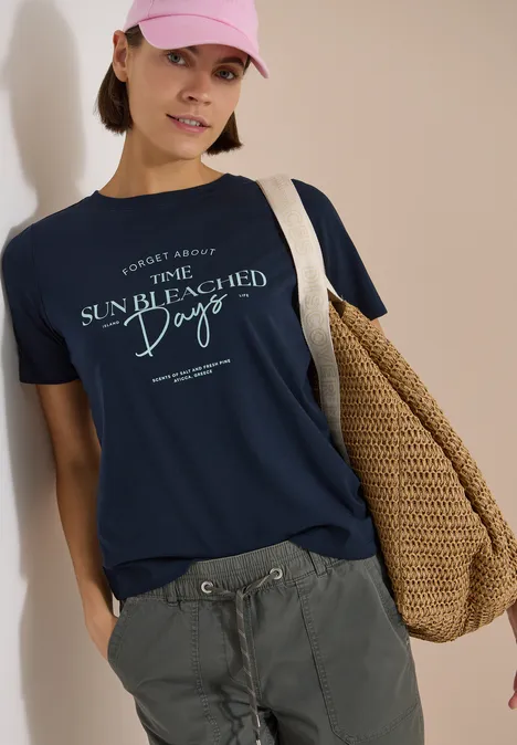 Shirt met boothals en Wording Artwork universal blue Persoon met marineblauw t-shirt “Sun Bleached Days”, grijze cargobroek en strooien tas.