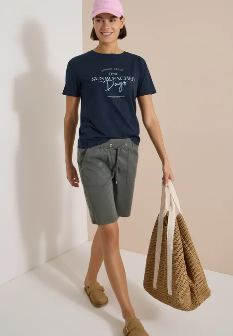 Shirt met boothals en Wording Artwork universal blue Een persoon draagt een navy T-shirt, grijze short, bruine mocassins en heeft een grote gevlochten tas bij zich.