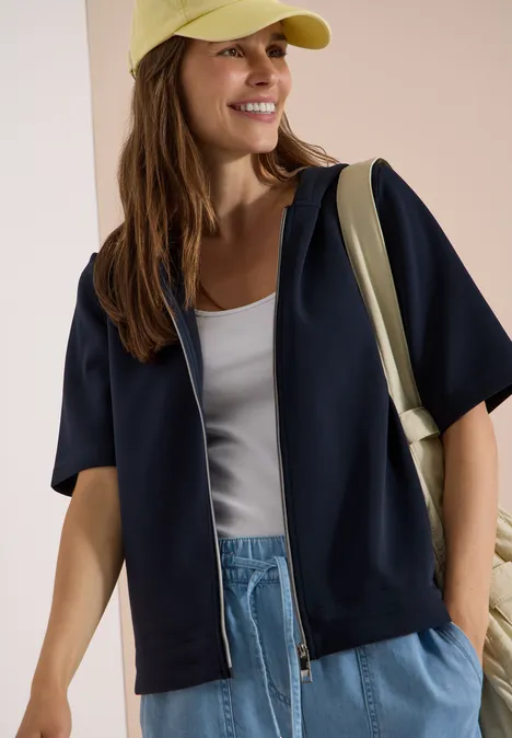 Frau in schwarzer Kapuzenjacke, weißem Top, blauen Jeans und gelber Mütze mit Schultertasche.
