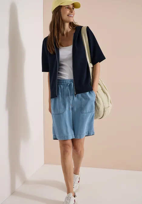 Frau im lässigen Outfit mit Jeans-Shorts, weißem Top, blauer Jacke, Sneakern, Cap und Tasche vor beiger Wand.
