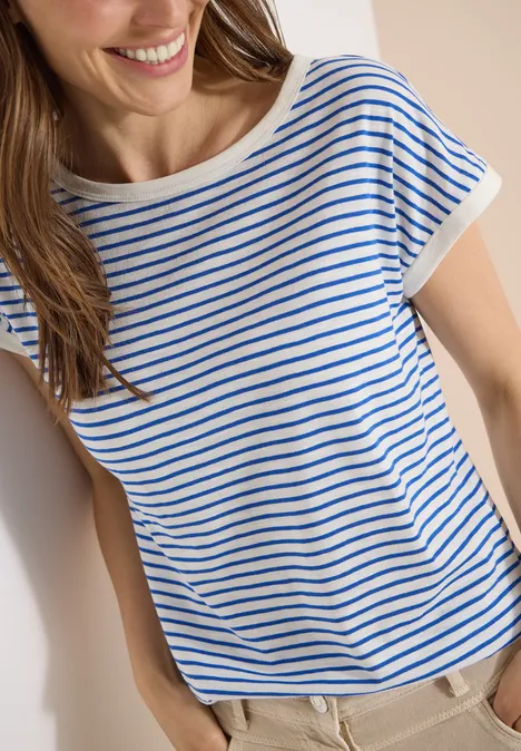 T-Shirt mit Boat Neck und Streifen surf blue Eine lächelnde Frau lehnt an der Wand, trägt ein blau-weiß gestreiftes T-Shirt und helle Hose.