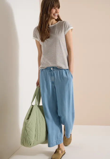 T-Shirt mit Boat Neck und Streifen urban taupe Eine lächelnde Frau trägt ein gestreiftes T-Shirt, weite blaue Hosen und eine grüne gesteppte Tasche.
