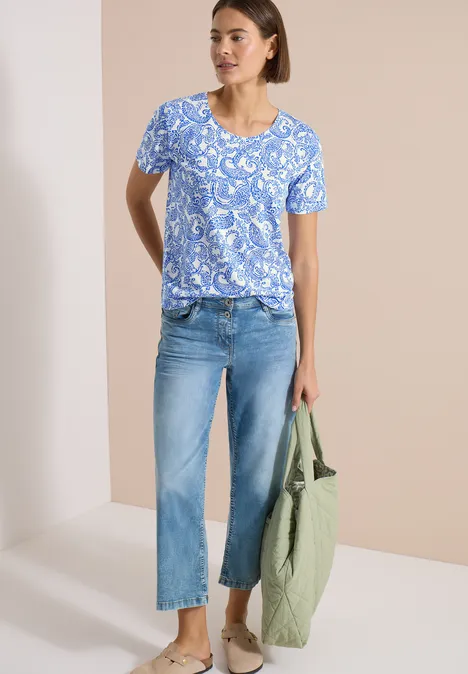 T-Shirt mit Rundhals und Muster vanilla white Frau trägt blau gemustertes T-Shirt, helle Jeans, grüne Tasche, Sandalen vor beigem Hintergrund.