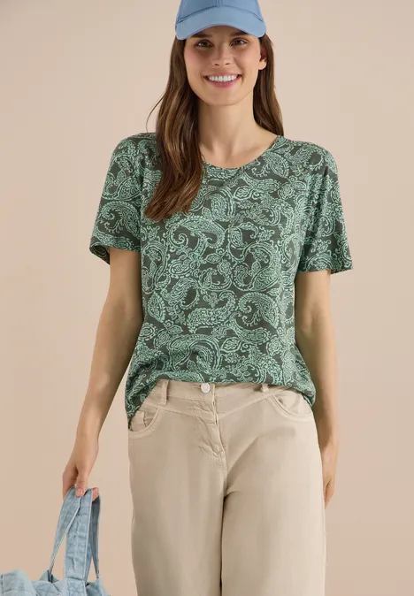 Frau trägt grünes, gemustertes T-Shirt, helle Hose und hält eine hellblaue Tasche.