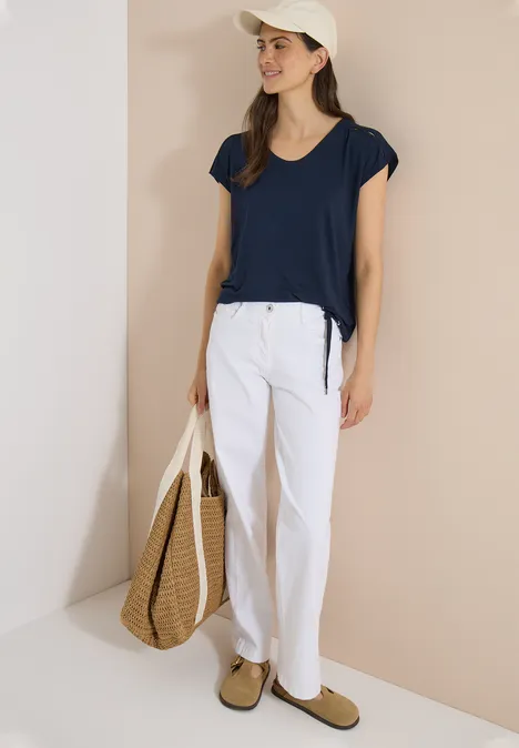 T-shirt à épaules tombantes avec découpe universal blue Femme en t-shirt bleu marine, pantalon blanc, casquette blanche, sac en paille et mocassins beige devant un mur beige.