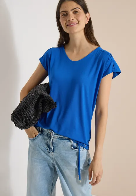 T-shirt à épaules tombantes avec découpe surf blue Femme portant un T-shirt bleu à cordon, jean clair et sac noir texturé à l’épaule, fond uni clair.