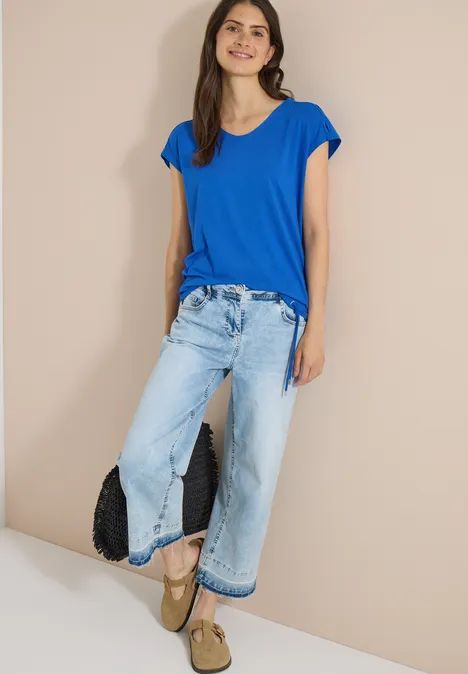 T-shirt à épaules tombantes avec découpe surf blue Femme en T-shirt bleu, jean large, sandales à plateforme et pochette noire devant un mur beige.