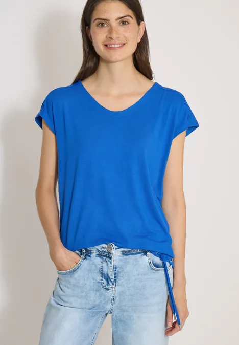 T-shirt à épaules tombantes avec découpe surf blue Femme en t-shirt bleu à col en V et jean clair, mains dans les poches, fond clair.