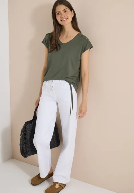 Shirt met afhangende schouders en uitgesneden detail khaki Vrouw in donkergroen T-shirt, witte jeans en bruine slippers met zwarte schoudertas voor een lichte muur.