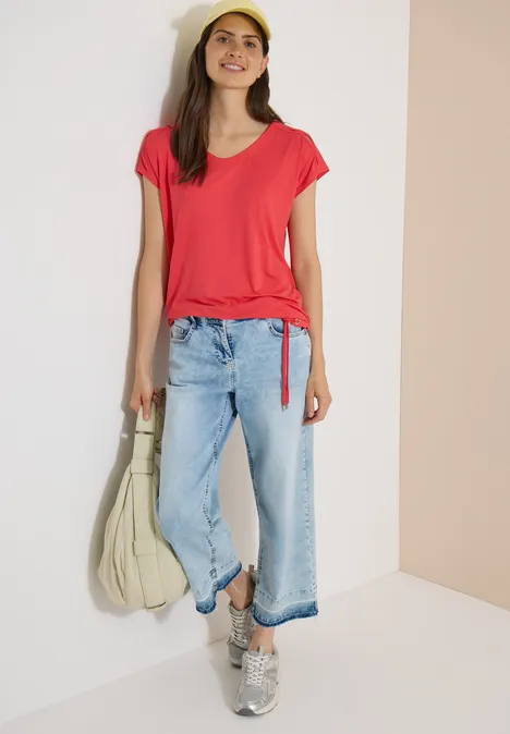 T-shirt à épaules tombantes avec découpe coral Femme en t-shirt rouge, jean clair et baskets, portant un grand sac, sur un fond neutre.