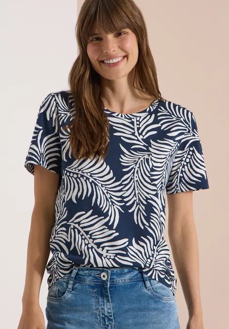 Frau trägt dunkelblaues T-Shirt mit weißem Blattmuster und Jeans, Gesicht unscharf.