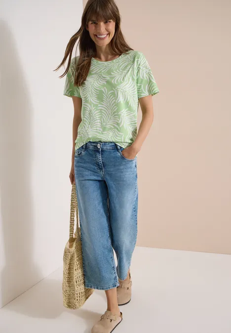Frau in grün-weiß gemustertem T-Shirt, weiten Jeans-Culottes, Basttasche und Sandalen.