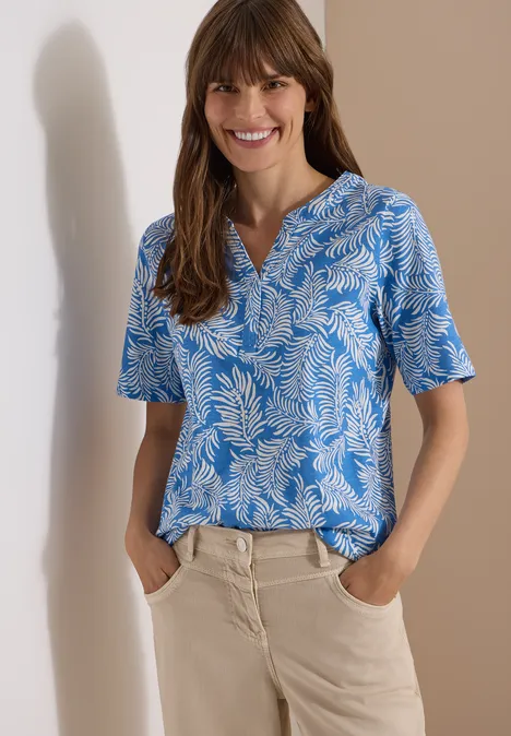Shirt met gespleten hals en print ethereal blue Vrouw in blauw-wit gebloemd shirt met korte mouw en lichte broek, handen in de zakken, voor beige achtergrond.