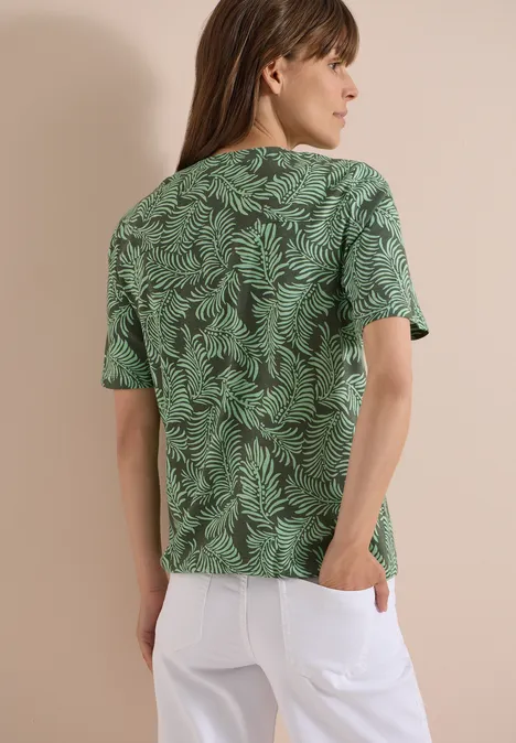 Shirt met gespleten hals en print khaki Achteraanzicht van een persoon met lang haar draagt een groen T-shirt met bladprint en een witte broek.