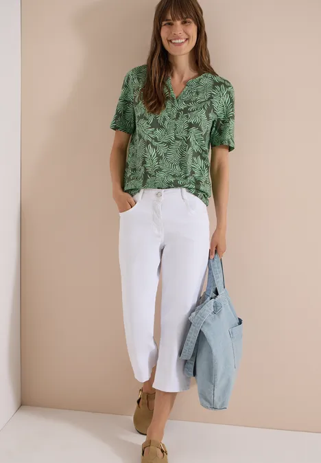 Shirt met gespleten hals en print khaki Vrouw met groene gebladerde top, witte capribroek en lichtblauwe spijkerjas over de arm