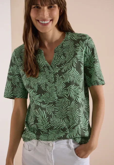 Shirt met gespleten hals en print khaki Vrouw met groen bladprintshirt en een witte broek, handen in de zakken.