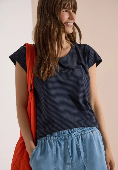 Frau trägt lockeres dunkelblaues T-Shirt, hellblaue Hose und rote Umhängetasche.