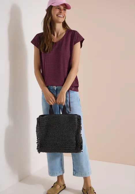 Vrouw in paarse top en spijkerbroek staat met zwarte tas in een lichte ruimte.