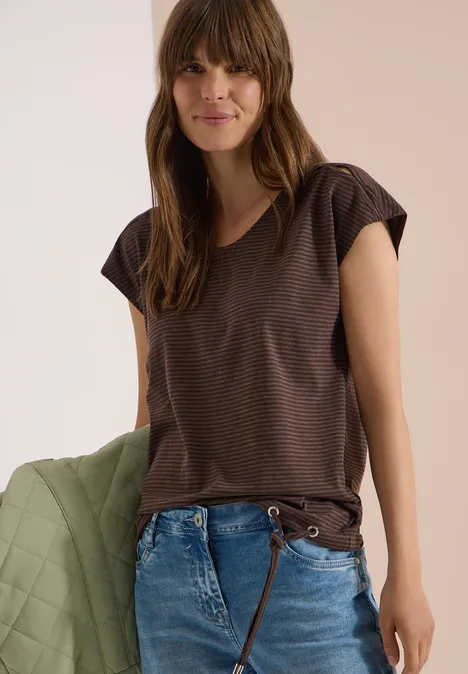 Vrouw draagt een bruin gestreept t-shirt met korte mouwen, blauwe jeans en houdt een groene doorgestikte jas vast.