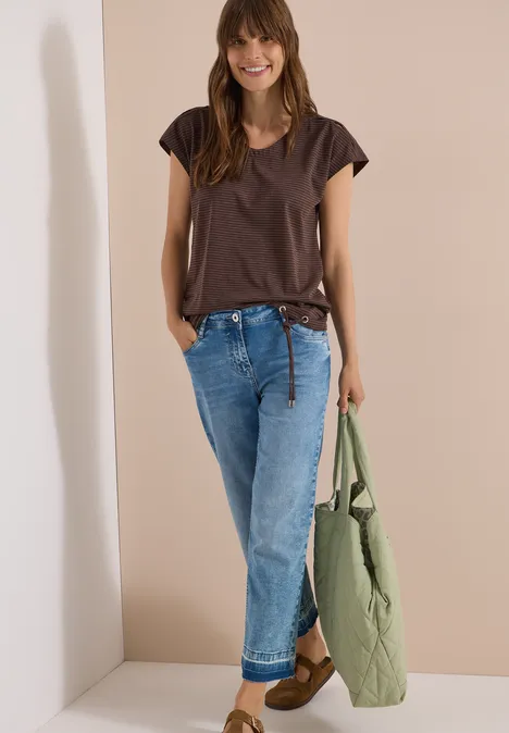 Vrouw voor lichte achtergrond, draagt bruin T-shirt, blauwe jeans en houdt groene gewatteerde jas vast.