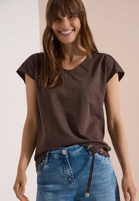 Vrouw in een bruin T-shirt met korte mouwen en een blauwe spijkerbroek voor een lichte achtergrond.