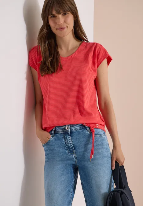 Frau in rotem T-Shirt mit Knoten, blauen Jeans, hält dunkle Tasche, steht vor hellem Hintergrund.