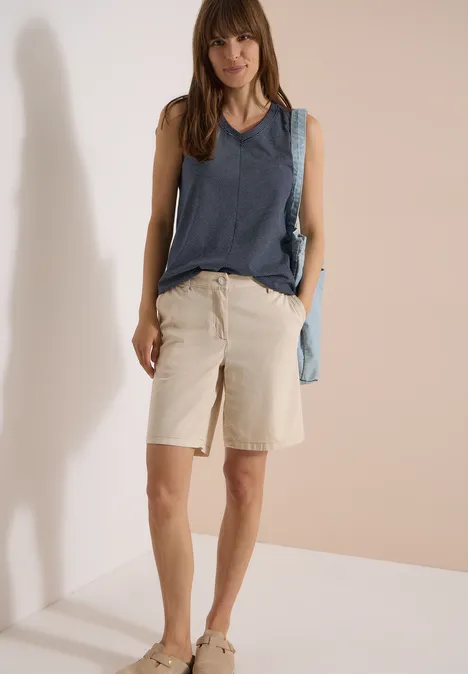 Top mit V-Neck und Streifen universal blue Frau mit ärmellosem Jeans-Top, beigen Bermudashorts, Sneakern und hellblauer Umhängetasche vor neutralem Hintergrund.