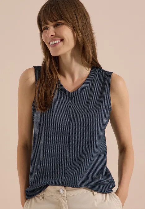 Top mit V-Neck und Streifen universal blue Frau trägt dunkelblaues ärmelloses Top mit V-Ausschnitt, Hände in den Taschen, vor beigem Hintergrund.
