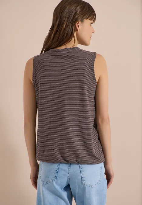Top met V-hals en strepen brown cacao Jonge vrouw van achteren, draagt een donkergrijs hemdje en lichtblauwe jeans, voor een beige achtergrond.