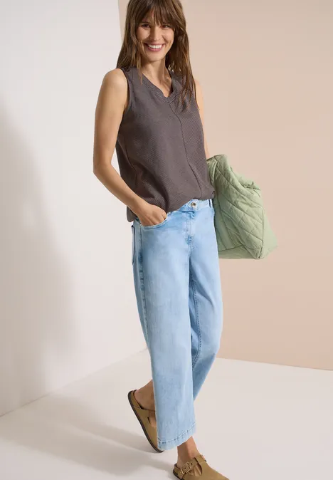 Top met V-hals en strepen brown cacao Vrouw in losse wijde jeans en mouwloos shirt, met een grote groene gewatteerde tas.