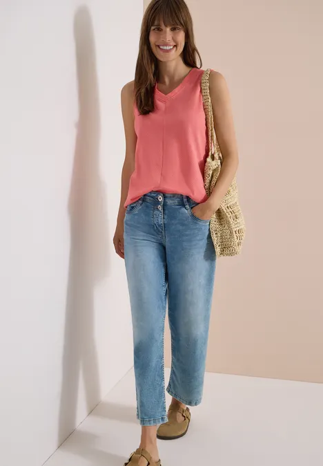 Top mit V-Neck und Streifen coral Frau in rosa ärmellosem Top, blauen Jeans, Basttasche und beigen Schuhen, stehend vor neutralem Hintergrund.