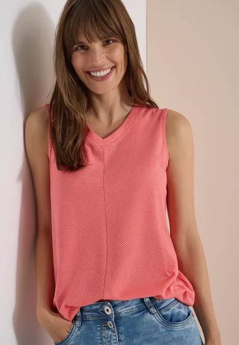 Top mit V-Neck und Streifen coral Frau im korallenfarbenen V-Ausschnitt-Top und Jeans lehnt entspannt an heller Wand.