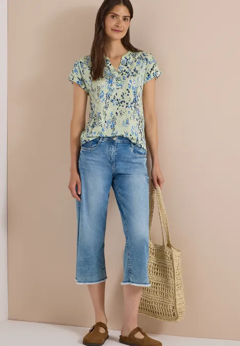 Frau in geblümter Bluse, blauen Jeans-Capripants und Strohtasche vor neutralem Hintergrund.
