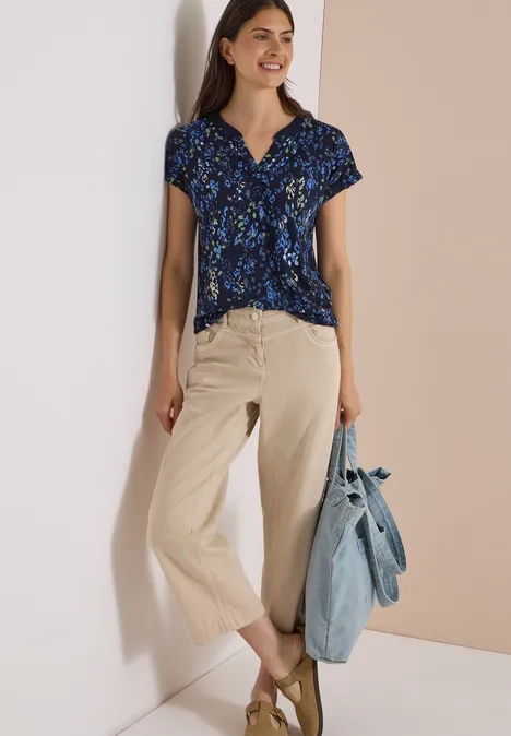 Frau in kurzärmliger dunkelblauer Bluse mit Blumenmuster, beiger weiter Hose und Jeans-Tasche.