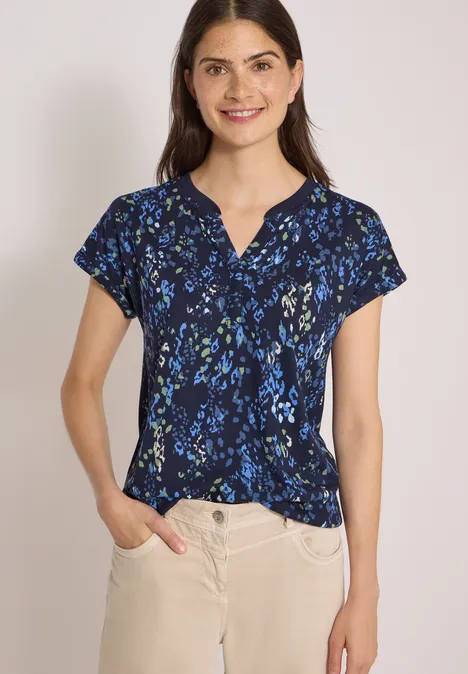 Frau mit marineblauer, kurzärmeliger Bluse mit Blumenmuster und beiger Hose vor hellem Hintergrund.