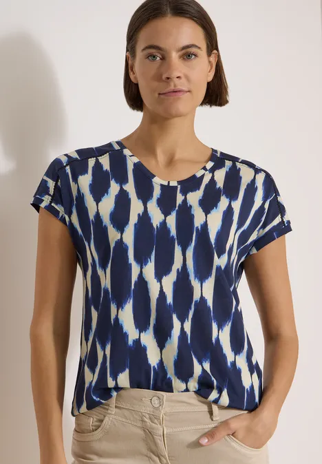 T-Shirt mit V-Neck und Spitzendetail urban dark blue Frau in kurzärmligem Shirt mit blau-weißem abstraktem Muster, eine Hand in Tasche, weißer Hintergrund.
