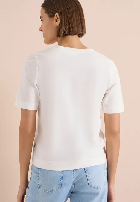 Frau mit kurzem Bob, weißem T-Shirt und hellen Jeans, von hinten vor beiger Wand stehend.