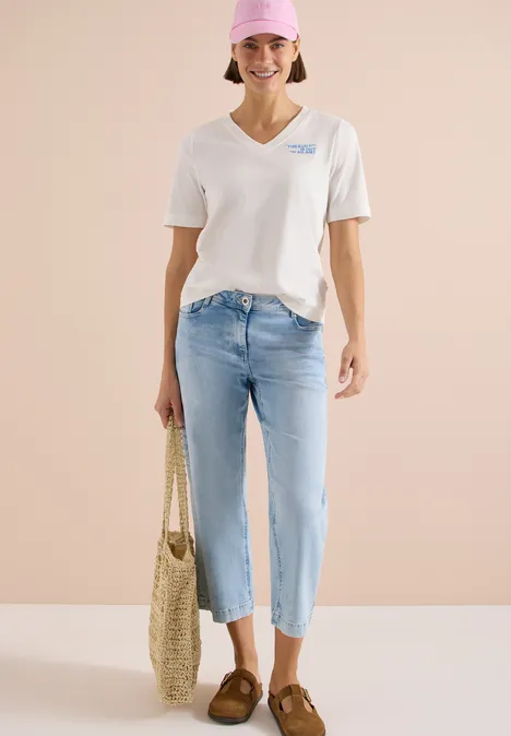 Frau in weißem V-Neck-Shirt, hellblauen Cropped-Jeans, braunen Loafers und Strohtasche vor beigem Hintergrund.