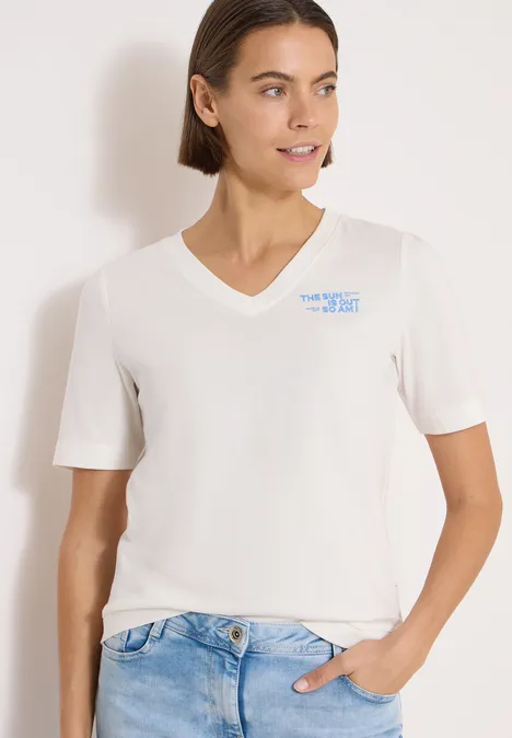 Frau in weißem V-Shirt und Jeans, eine Hand in der Hosentasche, vor hellem Hintergrund.