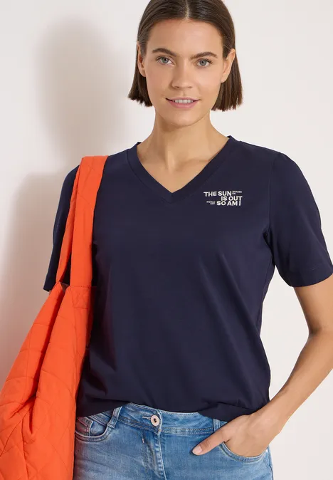 Frau trägt navy T-Shirt mit V-Ausschnitt, weiße Logo-Aufschrift, Jeans und orangene Umhängetasche.