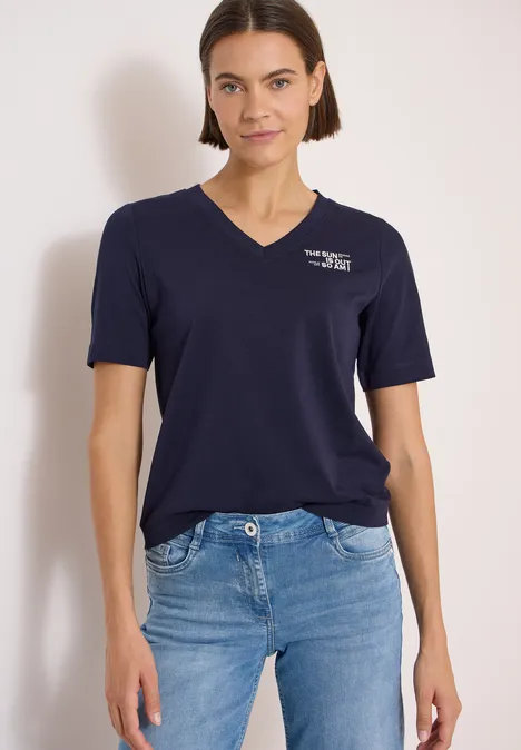 Frau in weißem T-Shirt und blauen Jeans vor hellem Hintergrund.